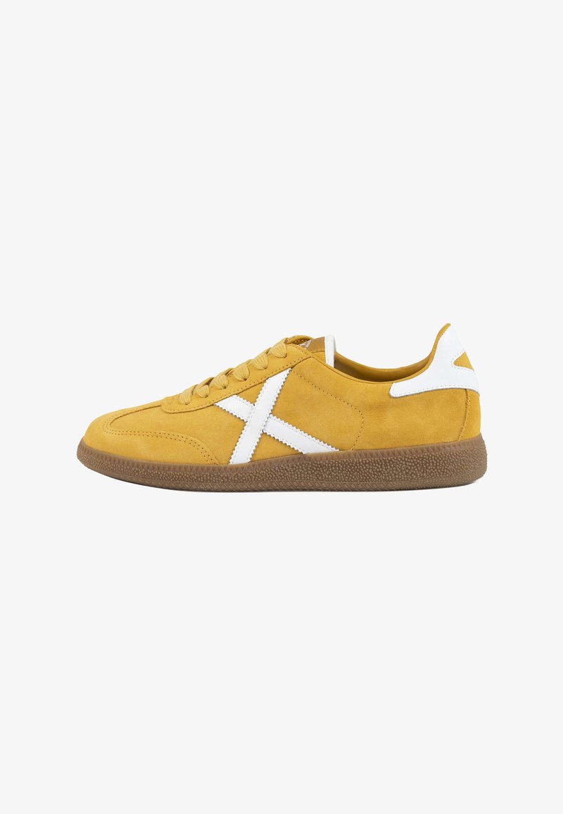 Baskets en daim jaune avec logo X en cuir blanc, semelle texturée marron et patch blanc au talon, présentées de profil sur fond blanc.