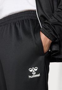 Svarta sportbyxor med elastisk midja, sidofickor och vitt "hummel"-logotyp. Materialet verkar slätt med en lätt textur.
