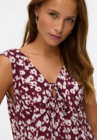 Femme aux longs cheveux bruns portant des boucles d'oreilles en perles et une robe sans manches à motifs bordeaux et blancs avec un détail de devant noué.