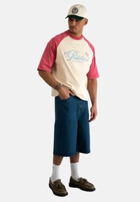T-shirt raglan à manches courtes crème et rose avec logo "Interdit", associé à un short en denim bleu et des chaussures marron. Chaussettes blanches visibles.