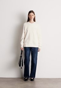 Κρέμα φούτερ με στρογγυλή λαιμόκοψη, συνδυασμένο με σκούρο μπλε παντελόνι σε φαρδιά γραμμή. Μαύρα παπούτσια χωρίς κορδόνια και μια μαύρη τσάντα με λουράκι. 