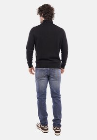 Maglione nero lavorato a maglia con colletto alto, indossato con jeans in denim azzurro chiaro e sneakers marroni con un logo bianco. Vista da dietro.