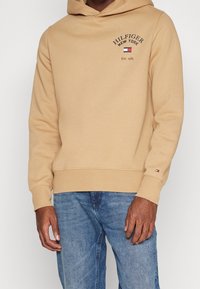 Felpa beige con cappuccio regolabile, tasca a marsupio e logo ricamato "HILFIGER NEW YORK". Tessuto morbido con polsini e orlo a coste.
