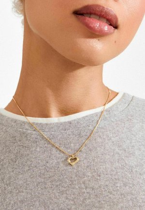 Zelta sirds formas kulons uz smalka ķēdītes, valkāts virs pelēka džempera ar baltu apkakli. Kulonam ir cilpu dizains.