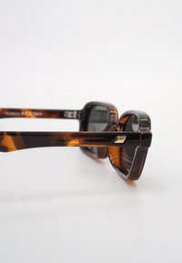 Καφέ ορθογώνια γυαλιά ηλίου σε σχέδιο χελώνας με σκοτεινούς φακούς που ακουμ πούν σε λευκή επιφάνεια, πλαϊνή όψη που δείχνει υφασμένα μπράτσα και μικρή μεταλλική λεπτομέρεια.