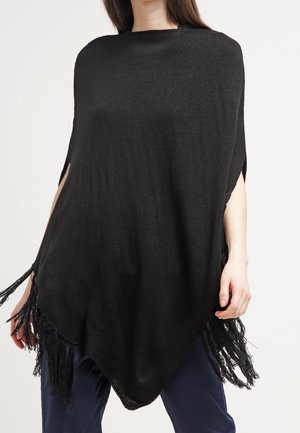 Vrouw draagt een losse zwarte gebreide poncho met franjes over een donkere broek, staand tegen een eenvoudige lichte achtergrond.