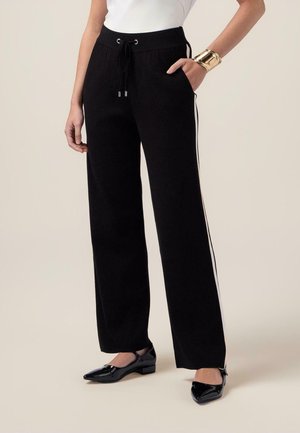 Kvinde iført sorte bukser med brede ben og hvide sidestriber, sorte spidse flade sko, hvid top og guldbandet armbånd, hånd i lommen.