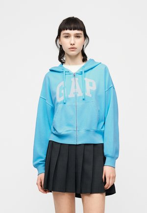 Jeune personne portant un sweat à capuche à fermeture éclair bleu clair de la marque GAP et une jupe plissée noire, debout devant un fond blanc uni.