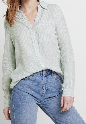 Frau trägt ein hellgrünes, gestreiftes Hemd mit Knöpfen, das in hoch taillierte blaue Jeans gesteckt ist, und steht vor einem einfarbigen Hintergrund.