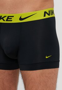 Czarne bokserki z szerokim neonowo-żółtym pasem w talii z nadrukiem "NIKE" w kolorze czarnym. Materiał jest gładki i dobrze przylega do ciała.