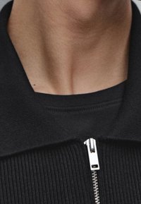 Schwarze Reißverschlussjacke mit gerippter Textur, ausgestattet mit einem silbernen Reißverschluss und einem Kragen. Wird über einem schwarzen Rundhalsausschnitt getragen.