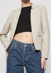 Beżowa, dopasowana marynarka o skróconym kroju, z gładką fakturą i dwoma kieszeniami na zamek, zestawiona z niebieskimi, wysokim stanem dżinsami.