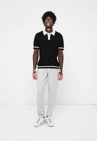 Fekete póló ing fehér gallérral és mandzsettával, szürke melegítőnadrággal és fehér sportcipővel párosítva. A modell egy egyszínű háttér előtt áll.