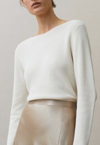 Vrouw in een crèmekleurige gebreide trui en een beige satijnen rok met een hoge taille, van schouders tot heupen afgebeeld tegen een neutrale achtergrond.