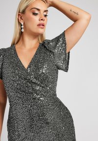 Hopeapintaisella paljeteilla koristeltu mekko, jossa on syvä V-kaula-aukko ja lyhyet puffihihat. Mekossa on istuva vyötärö ja kimalteleva, teksturoitu pinta.