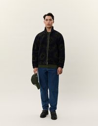 Ulllete marinejakke med grønt geometrisk mønster, glidelås foran, kombinert med en mørkegrønn t-skjorte, blå jeans og en grønn caps holdt i hånden.