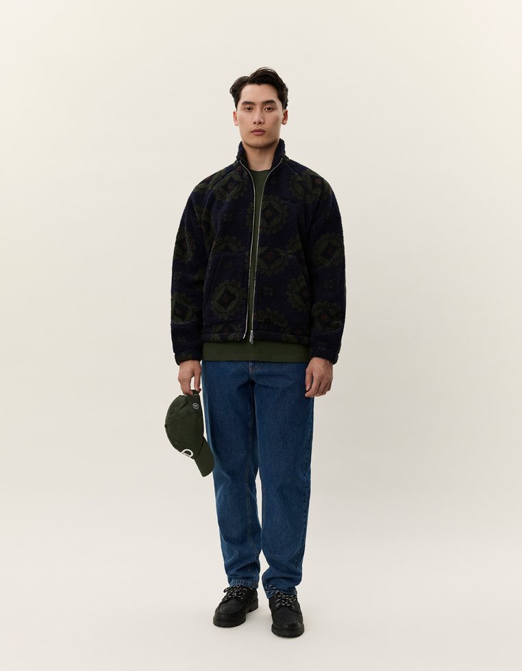 Ulllete marinejakke med grønt geometrisk mønster, glidelås foran, kombinert med en mørkegrønn t-skjorte, blå jeans og en grønn caps holdt i hånden.