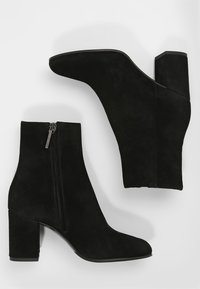 Bottines en suede noir avec un talon carré, bout pointu, détail de fermeture éclair sur le côté et une texture lisse, mettant en avant un design épuré et moderne.