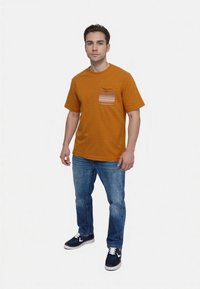 Camiseta a rayas naranja con un bolsillo a rayas horizontales, combinada con jeans azules y zapatillas marinas con acentos blancos.