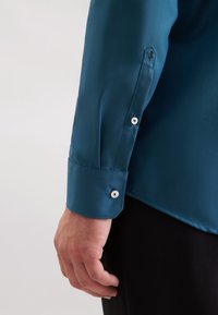 Der linke Arm eines Mannes in einem türkisfarbenen Langarmshirt mit weißen Knöpfen und bestickten Details an der Manschette, der neben schwarzen Hosen ruht.