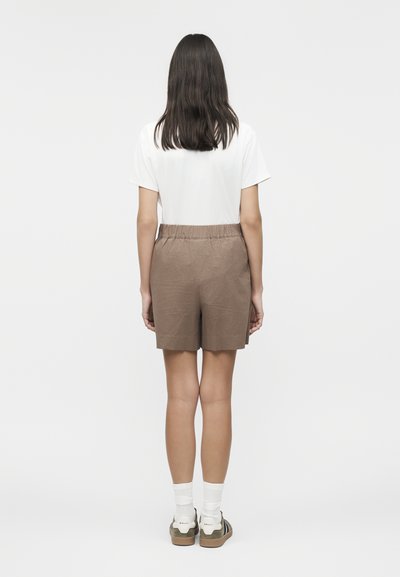 Frau steht mit dem Rücken zur Kamera, trägt ein weißes kurzärmliges Hemd, braune, strukturierte Shorts, weiße Socken und grüne Turnschuhe.