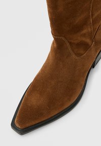 Καφέ σουέντ μποτάκι μέχρι τον αστράγαλο με μυτερή μύτη και μαύρη σόλα, με ορατές ραφές στο πάνω μέρος.