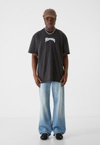 Sort oversized t-shirt med hvid grafisk print, løse lyseblå denimjeans og sorte sneakers. Brun kasket og kædedesign inkluderet.