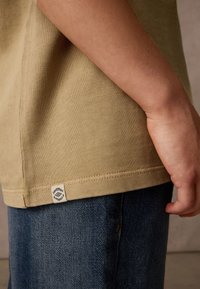 Beige ribbad t-shirt med en tätt sittande silhuett, med en logotypetikett vid fållen. Bärs tillsammans med blå denimjeans.