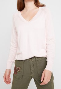 Světle růžový svetřik s dlouhými rukávy a výstřihem do V, s žebrovanými manžetami, v kombinaci s olivově zelenými kalhotami se květinovou výšivkou na levé kapse.