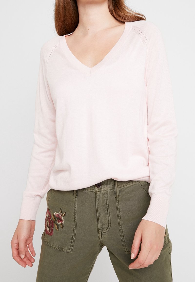 Světle růžový svetřik s dlouhými rukávy a výstřihem do V, s žebrovanými manžetami, v kombinaci s olivově zelenými kalhotami se květinovou výšivkou na levé kapse.