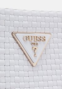 Surface tissée blanche texturée avec un logo triangulaire Guess USA doré et blanc comportant un point d'interrogation au centre.