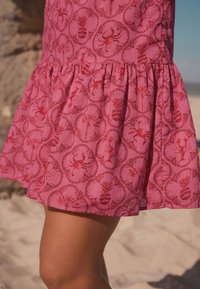 Vestito di tessuto rosa con motivo di granchi e ananas, orlo svasato e dettagli con vita arricciata. Tessuto liscio e leggero.