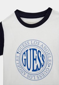 T-shirt bianco in cotone con scollatura e maniche a costine blu navy. Presenta un grande logo circolare blu con il testo "GUESS LOS ANGELES" al centro.