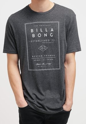 Mann trägt ein dunkelgraues Billabong-T-Shirt mit weißem rechteckigem Logo und dem Text "Established '73" sowie mit Händen in den Taschen.
