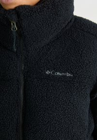 Närbild av en svart fleecejacka med en frontdragkedja och en broderad Columbia-logotyp på bröstet.