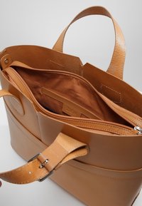 Bruine leren tote bag met dubbele handvatten, met een ruime binnenkant gevoerd met bruine stof; bevat een ritszak en een merketiket.