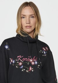 Haut à capuche noir avec une texture en chevrons ; présente une broderie florale en rouge, bleu et blanc. Comprend un col haut et des accents de cordon.