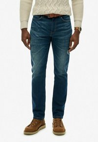 Hombre vistiendo jeans de mezclilla azules, cinturón de cuero marrón, suéter de punto trenzado beige, botas de cuero marrón con cordones y reloj de pulsera, de pie frente a un fondo blanco.