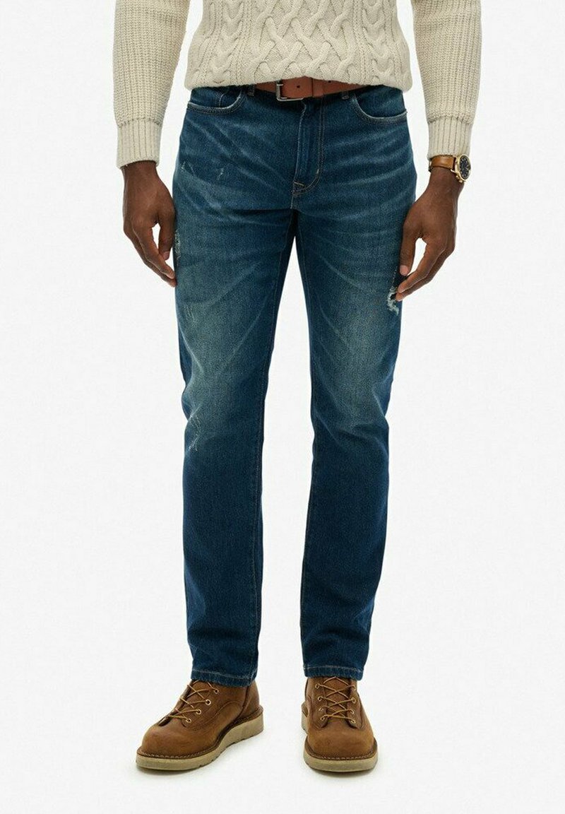 Hombre vistiendo jeans de mezclilla azules, cinturón de cuero marrón, suéter de punto trenzado beige, botas de cuero marrón con cordones y reloj de pulsera, de pie frente a un fondo blanco.