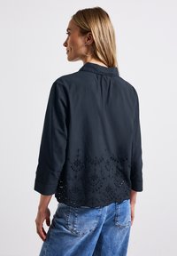 Camicia nera in cotone con colletto alla cinese, maniche a tre quarti e dettagli in pizzo ricamato all'orlo. Indossata con jeans in denim blu.