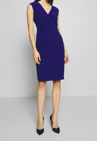 Lila, knälång klänning med djup V-ringning och omslagssdesign. Har ärmlös skärning och ett slätt, texturerat tyg.