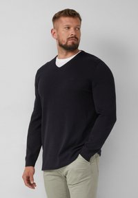 Marineblauer V-Ausschnitt-Pullover aus weichem Strickstoff, mit langen Ärmeln und gerippten Bündchen. Kombiniert mit hellen, eng geschnittenen Hosen.