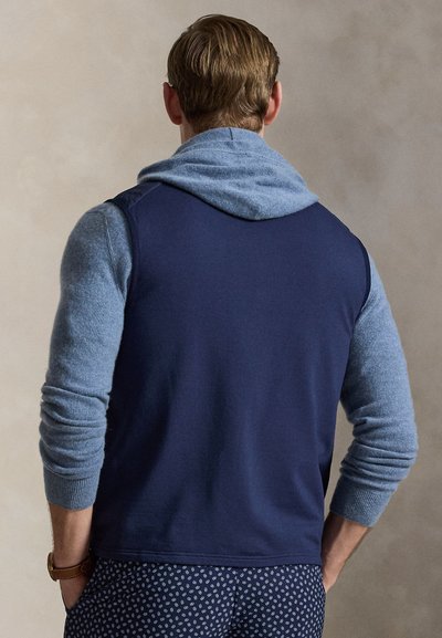 Άνδρας με κοντά ξανθά μαλλιά στέκεται με την πλάτη στραμμένη, φοράει μπλε γιλέκο πάνω από μπλε φούτερ με κουκούλα και σκούρο παντελόνι με σχέδια, με τα χέρια στις τσέπες.