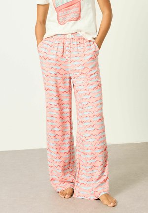 Osoba nosiaca voľné ružové pyžamové nohavice s modrými a červenými vlnitými pruhmi, stojaca na boso s rukami v vreckách.