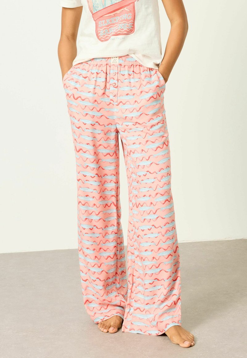 Personne portant un pantalon de pyjama rose ample avec des motifs de rayures ondulées bleues et rouges, debout pieds nus les mains dans les poches.