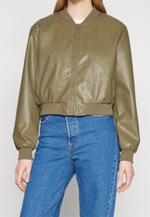 Kobieta w oliwkowej bomberce zapinanej na zamek z prążkowymi mankietami i pasem, w połączeniu z niebieskimi dżinsami z wysokim stanem.