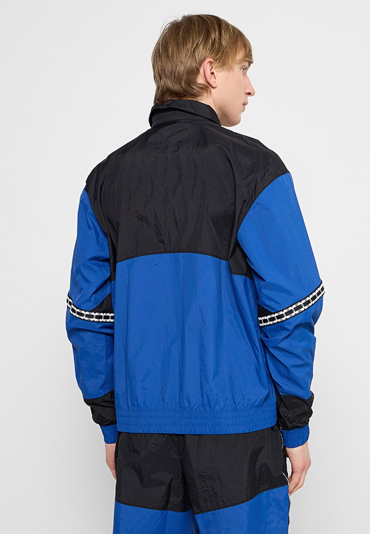 Chaqueta cortaviento azul y negra con cuello alto, puños elásticos y un acento con patrón a lo largo de las mangas. Tela ligera con una textura suave.