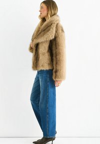 Faux fur coat in light brown, featuring a wide collar and loose fit, paired with high-waisted blue jeans and dark heeled boots.