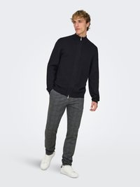 Uomo in piedi con le mani nelle tasche, indossa un maglione nero con cerniera, pantaloni grigi a quadri e sneakers bianche.