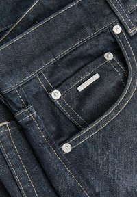Τσέπη από σκούρο μπλε τζιν με ορατές μπεζ ραφές, ασημένια καρφιά και μια μικρή μεταλλική ετικέτα με την επιγραφή «EST. 1990.»